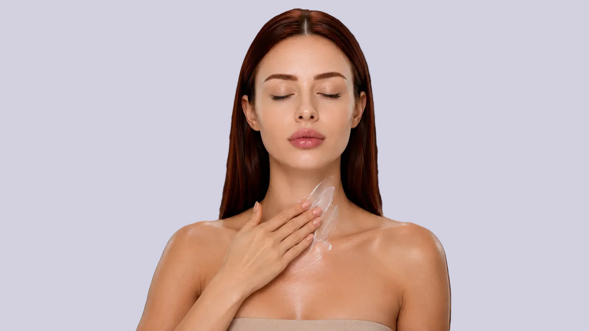 A woman with long dark red hair is shown from the chest up against a solid white background, applying a clear, transparent sheet mask to her neck and upper chest with her hand.
