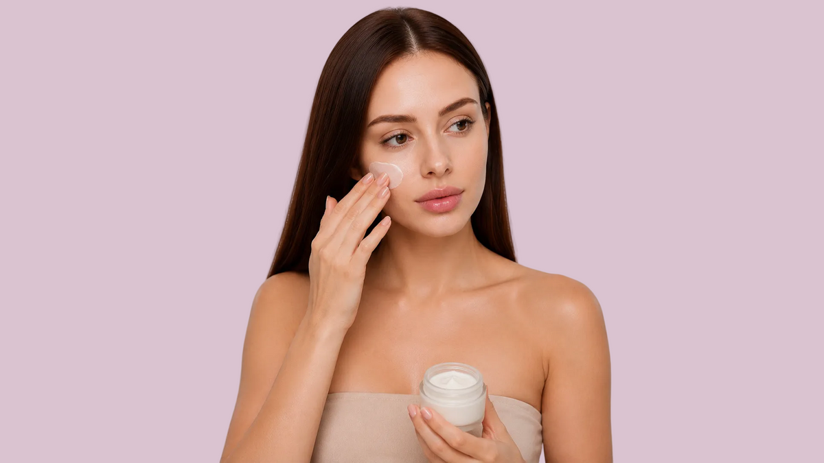 A woman with long dark hair applies a cream product to her cheek while holding a small white jar against a solid light purple background.