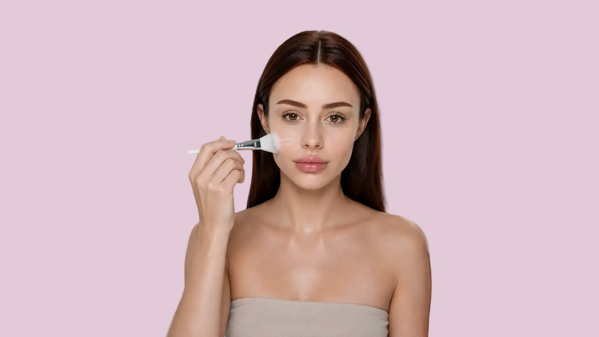 A woman with long brown hair is shown from the chest up against a light lavender background, holding a makeup brush and applying cream makeup to her cheek. She is wearing a beige strapless top.