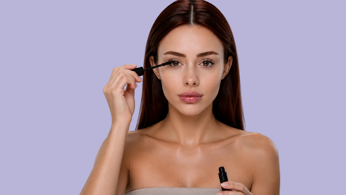 A woman with long dark brown hair applies mascara to her upper lashes while holding a black cosmetic tube in her other hand, set against a solid gray studio background.