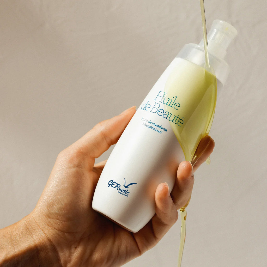 A hand holds a white bottle of QP Réalité 'Huile de Beauté' beauty oil, with golden-yellow oil being poured from the clear pump into a stream against a neutral beige background.