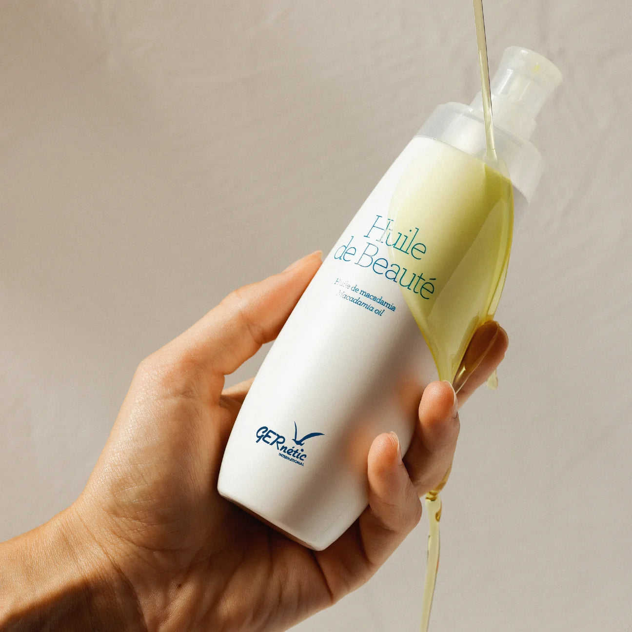 A hand holds a white bottle of QP Réalité 'Huile de Beauté' beauty oil, with golden-yellow oil being poured from the clear pump into a stream against a neutral beige background.