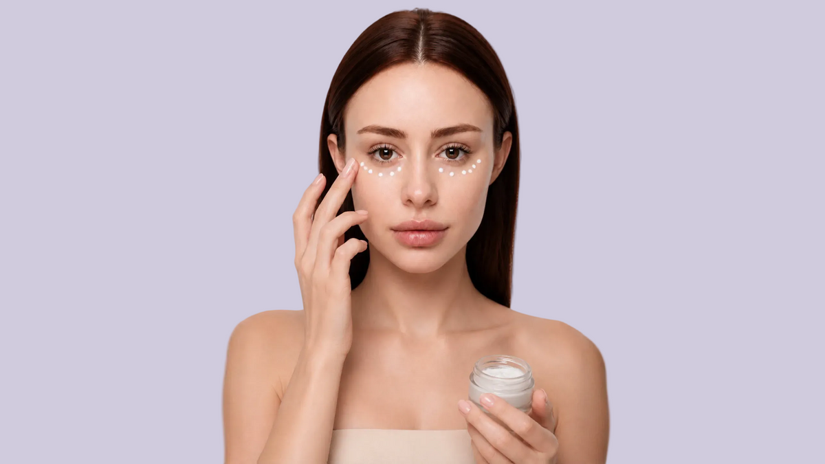 A woman with dark hair applies a skincare cream from a small clear jar to her cheek, with white dot makeup visible under her eyes, against a light gray background.