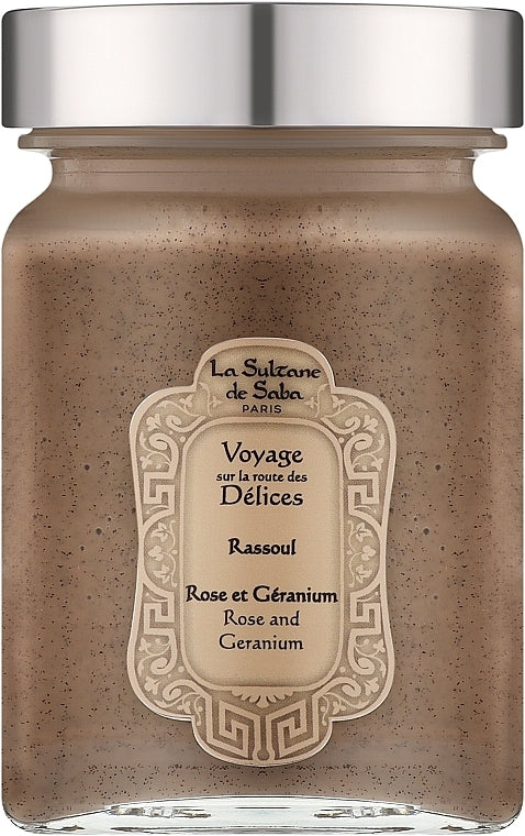 A jar of La Sultane de Saba Paris 'Rassoul' body scrub, featuring a speckled brownish-beige scrub inside a clear glass jar with a metallic silver lid and a cream-colored decorative label. Rassoul Face, Body & Hair Mask