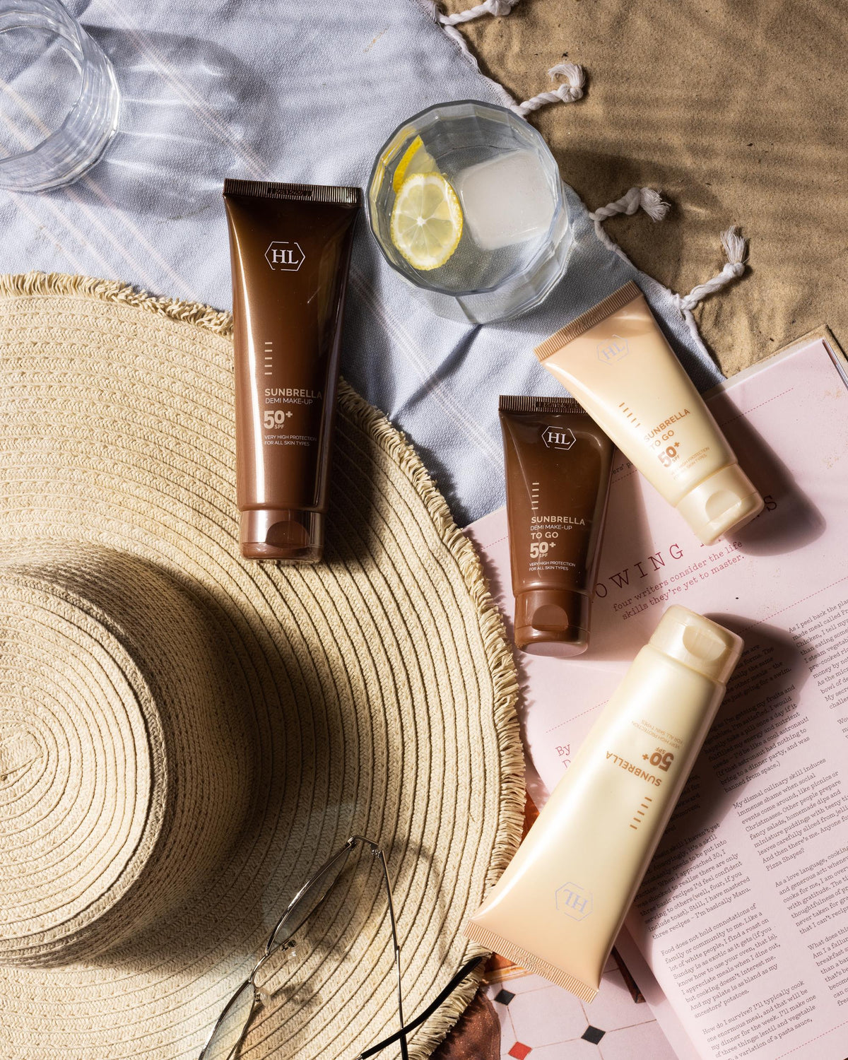 Four cosmetic tubes from the brand HL are arranged in a styled flat-lay with a straw hat, a glass of water with a lemon slice, and a magazine on a beach-themed blanket background.