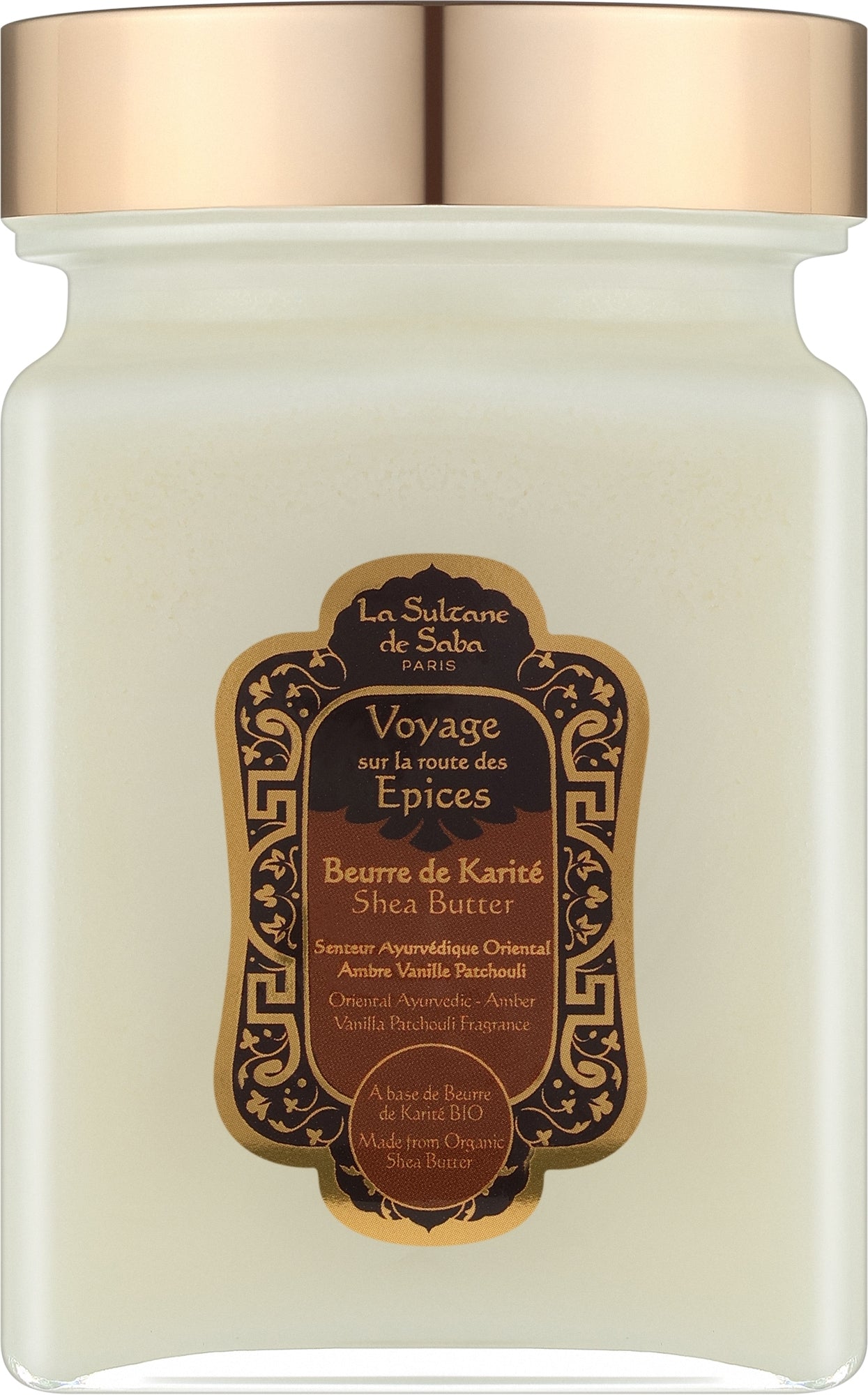 A jar candle with a frosted glass body showing a warm cream-colored wax, topped with a metallic rose gold lid. The front features an ornate gold and dark brown label for 'La Sultane de Saba' 'Voyage sur la route des Epices' (Shea Butter Spice Journey).