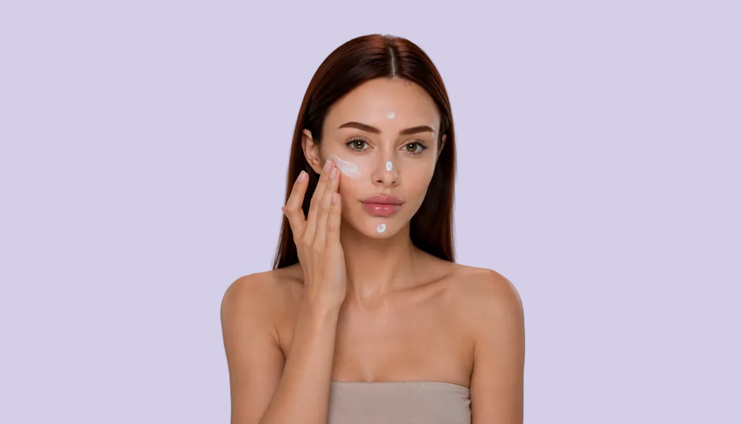 A woman with long dark brown hair applies a white cream or makeup to her cheek against a plain gray studio background.